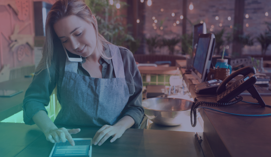 Una mujer que lleva un delantal habla por teléfono mientras utiliza una tableta en la barra de un restaurante; cerca de ella se ven un ordenador y otros equipos.