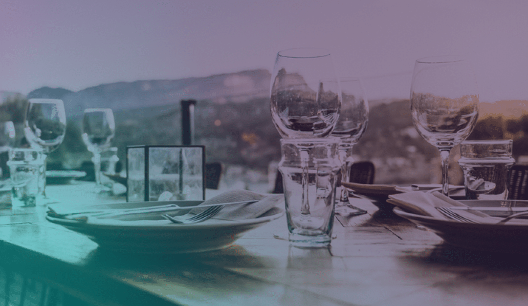 Elegante juego de mesa para comer al aire libre con copas de vino, platos y servilletas, con una vista panorámica de las montañas difuminada al fondo.