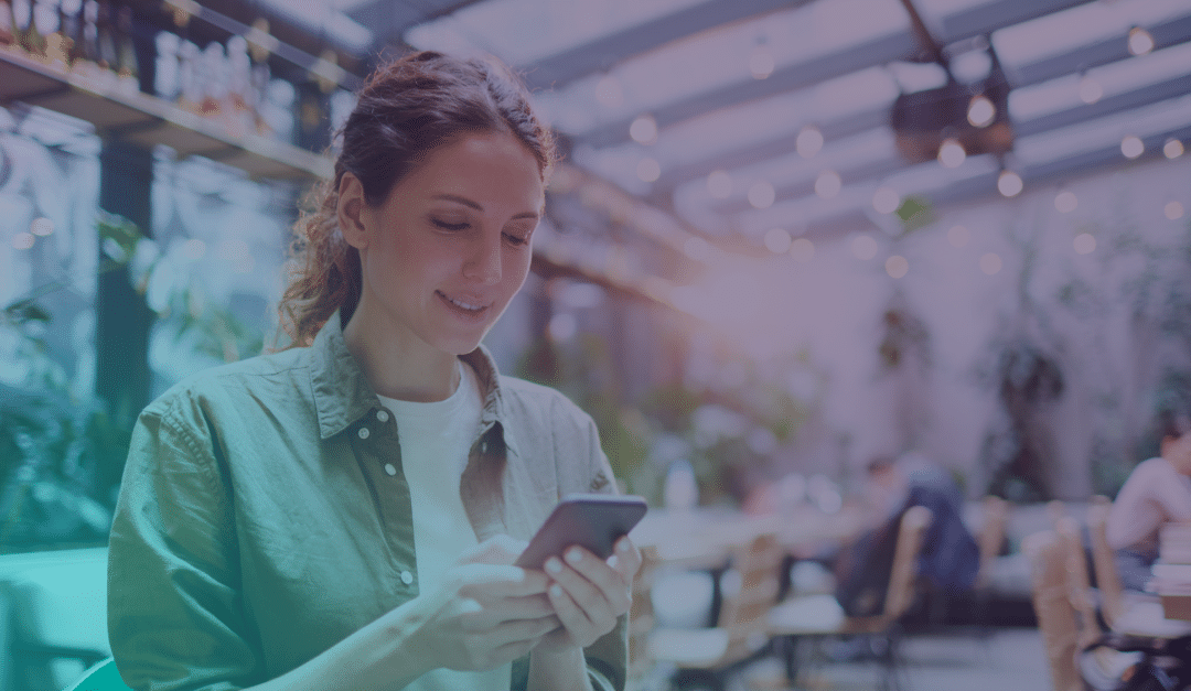 Una mujer de pelo castaño sonríe mientras utiliza su smartphone en una cafetería luminosa con lámparas colgantes y plantas. En el fondo se ven otras personas borrosas.
