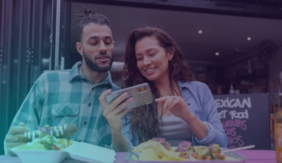 Un hombre y una mujer están sentados en una mesa al aire libre con comida, sonriendo y mirando juntos un teléfono móvil frente a un food truck con un cartel con el menú.