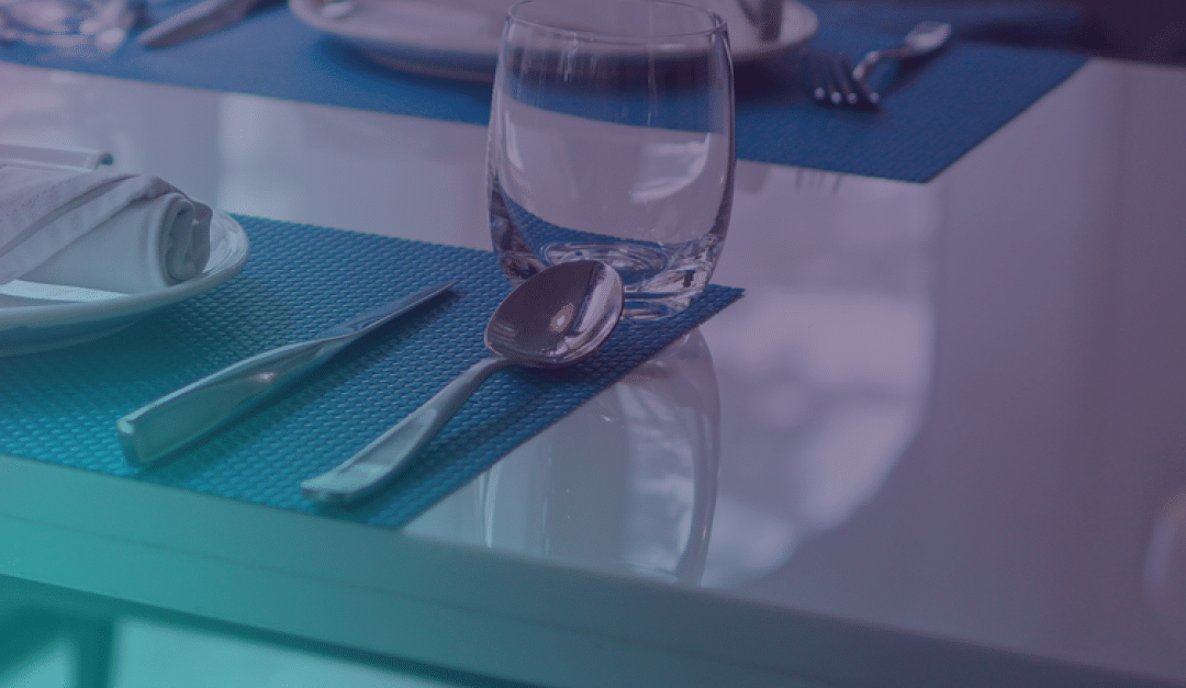 Un vaso, una cuchara y un cuchillo descansan sobre un mantel individual azul, junto a un plato blanco con una servilleta doblada, sobre una mesa brillante, puesta para la comida.