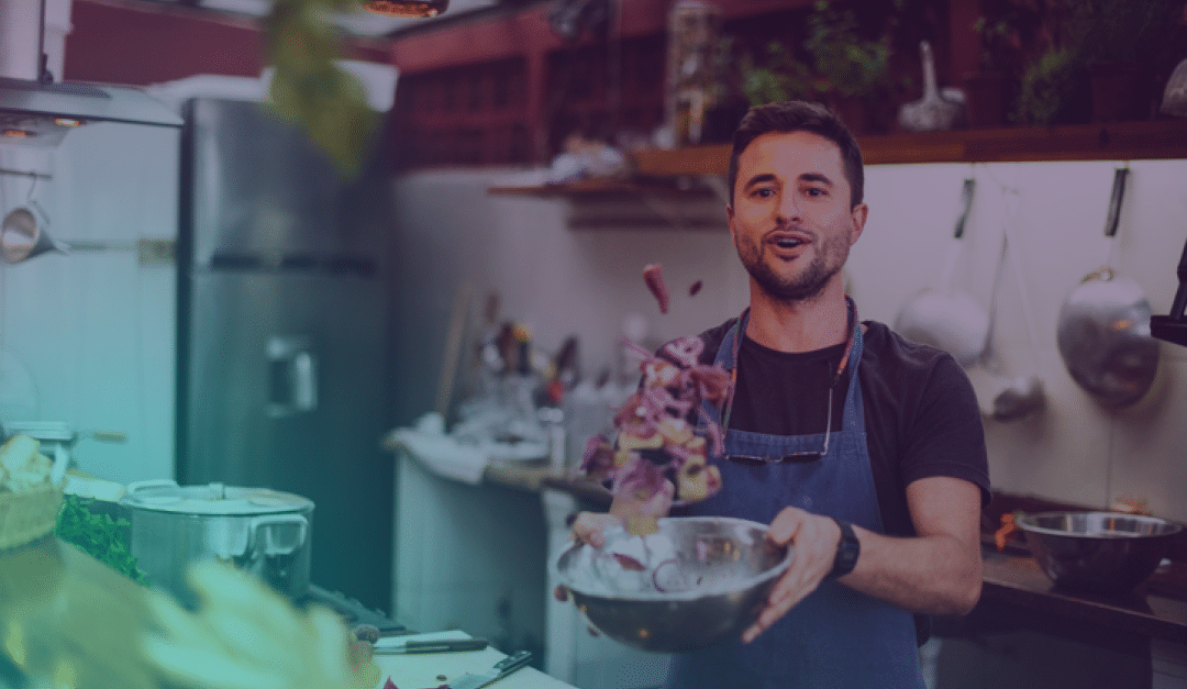 Un hombre en una cocina, con un delantal azul, está removiendo verduras en un cuenco de metal, sonriendo mientras cocina. Al fondo se ven ollas, utensilios y otros objetos de cocina.