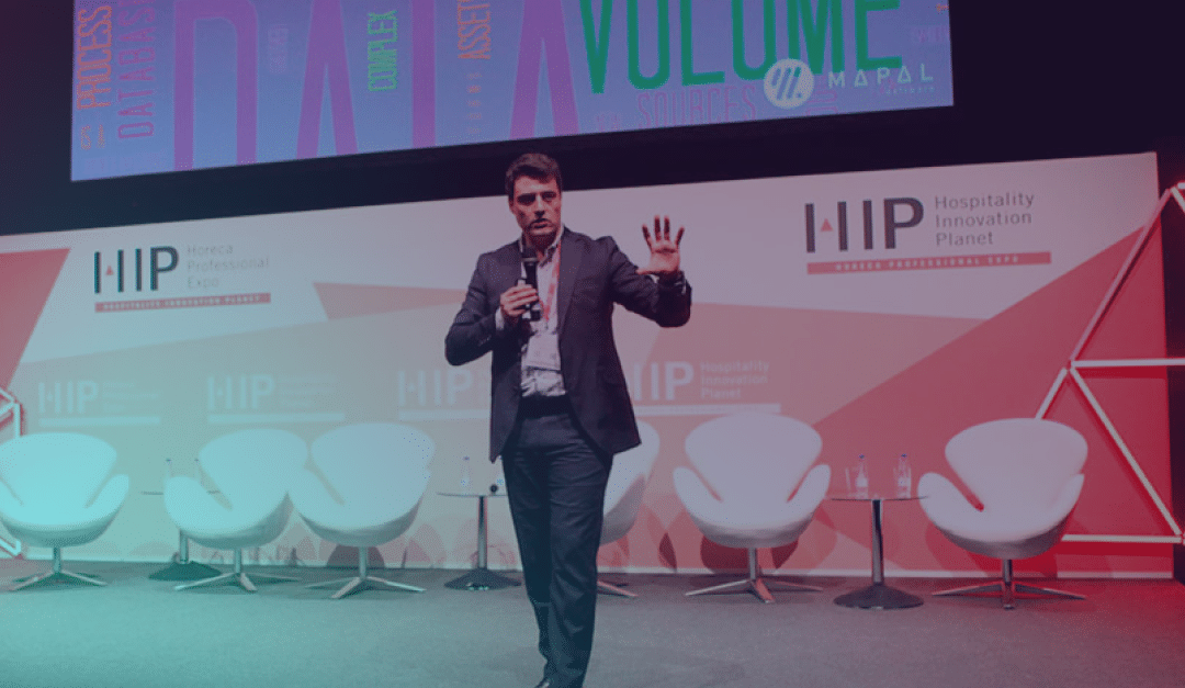 Un hombre vestido con traje habla en el escenario del HIP, haciendo gestos con una mano. Se ven cuatro sillas blancas y una botella de agua. Al fondo hay una pantalla de colores con texto y logotipos.