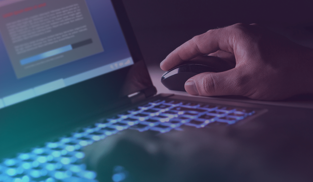 Una mano maneja el ratón de un ordenador junto a un portátil con el teclado iluminado y un mensaje de advertencia borroso en la pantalla, lo que sugiere un problema de ciberseguridad o una amenaza en línea.