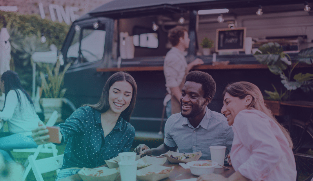 Tres personas sentadas en una mesa al aire libre, haciéndose un selfi y sonriendo, con comida y bebidas delante de ellas y un food truck negro al fondo.