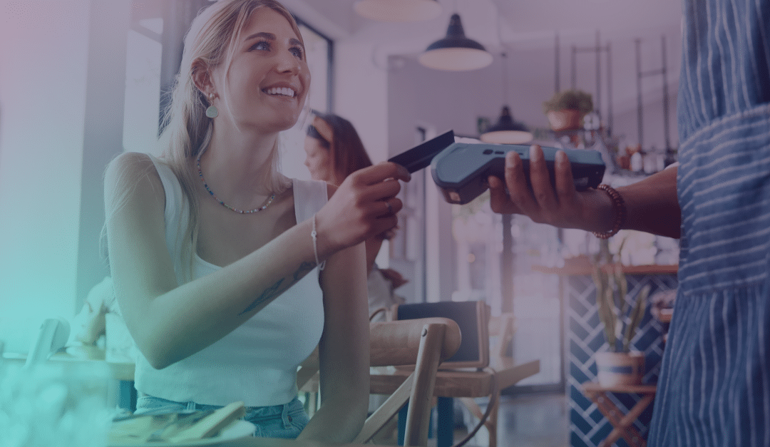 Una mujer sonriente, con una camiseta sin mangas blanca, está sentada en la mesa de una cafetería y sostiene un teléfono para acercarlo a un lector de tarjetas que tiene en la mano una persona que lleva delantal, lo que sugiere que está realizando un pago sin contacto.