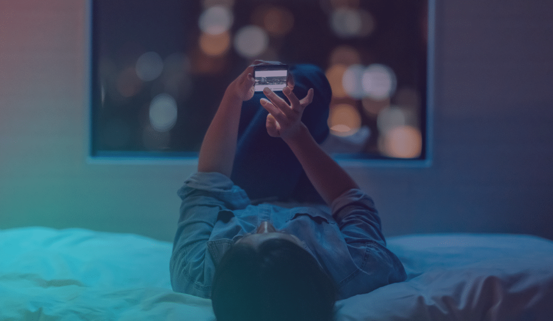 Una persona tumbada en la cama por la noche, mirando un smartphone, con las luces difusas de la ciudad visibles a través de una ventana al fondo.