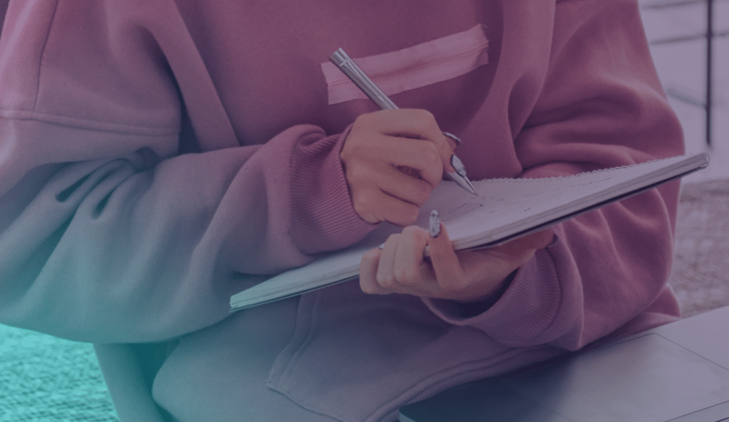 Una persona con una sudadera rosa escribe en un cuaderno con un bolígrafo plateado mientras está sentada en un interior. Solo se le ven las manos y el torso.