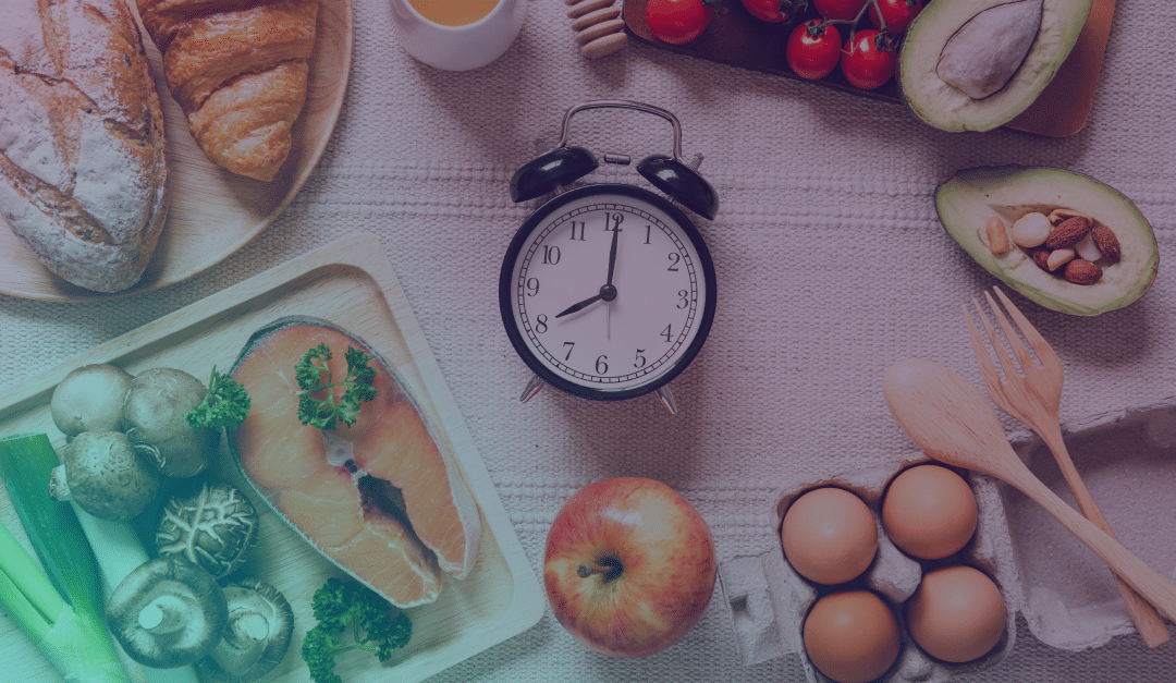 Un reloj rodeado de diversos alimentos, entre ellos salmón, huevos, pan, aguacate, setas, tomates, miel y una manzana, dispuestos sobre una mesa junto a utensilios de cocina.