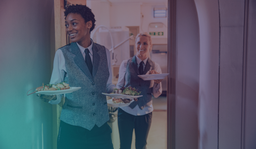 Dos camareros uniformados atraviesan una puerta con bandejas de comida, sonriendo mientras sirven las comidas. El escenario parece ser un restaurante o un evento de catering.