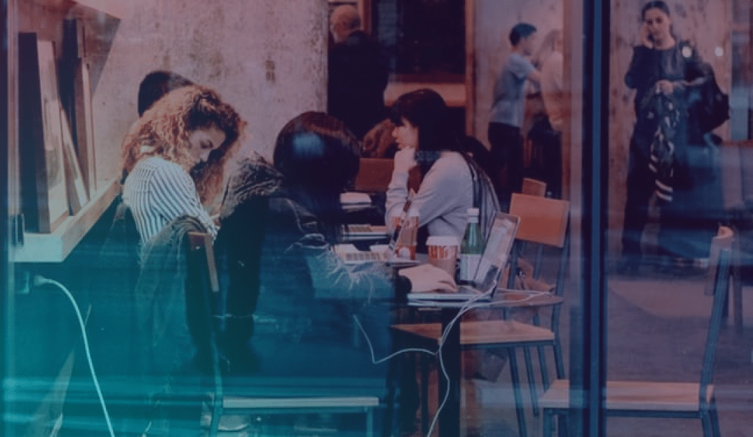 Hay gente sentada en una mesa dentro de una cafetería, trabajando con sus ordenadores portátiles y charlando. Se ven estanterías con libros y, a través de la ventana, se puede ver a los transeúntes que pasan por la calle.