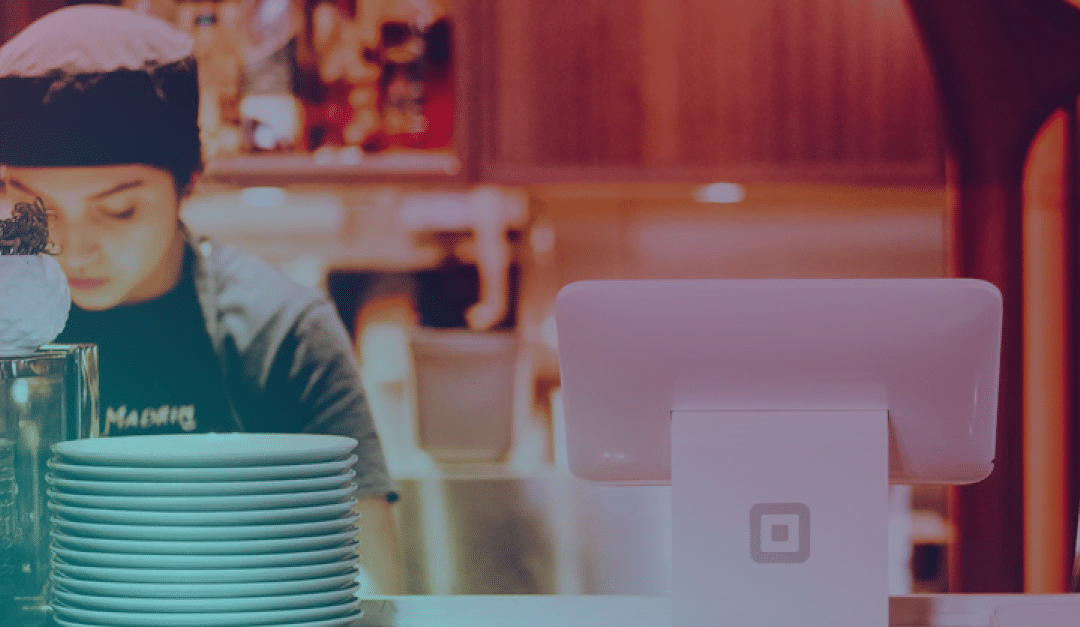Un empleado de un restaurante está de pie detrás de una pila de platos y una caja registradora digital, concentrado en su trabajo. Al fondo se ven una iluminación cálida y unas estanterías difuminadas.