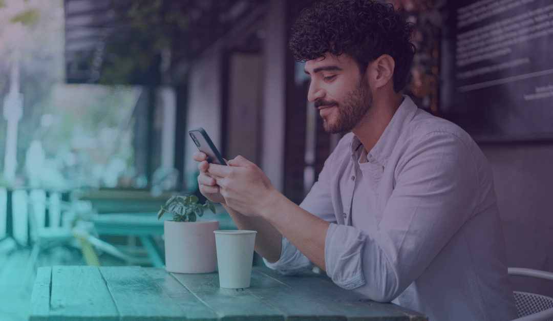 Un hombre con el pelo rizado está sentado en la mesa de una cafetería al aire libre, sonriendo mientras mira su smartphone. Delante de él hay una planta en una maceta y un vaso de papel.