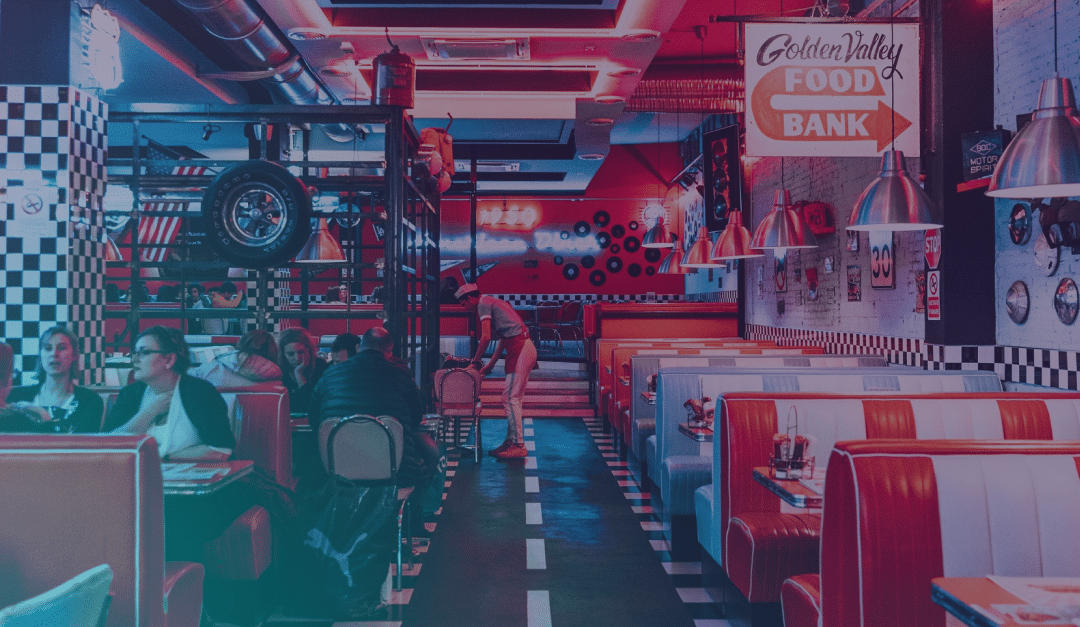 Interior de una cafetería de estilo retro con reservados rojos, paredes a cuadros, luces de neón y decoración vintage. Hay gente sentada en las mesas. Un cartel dice «Golden Valley Food Bank». Una persona está de pie cerca del fondo, junto a unos adornos con motivos de coches.