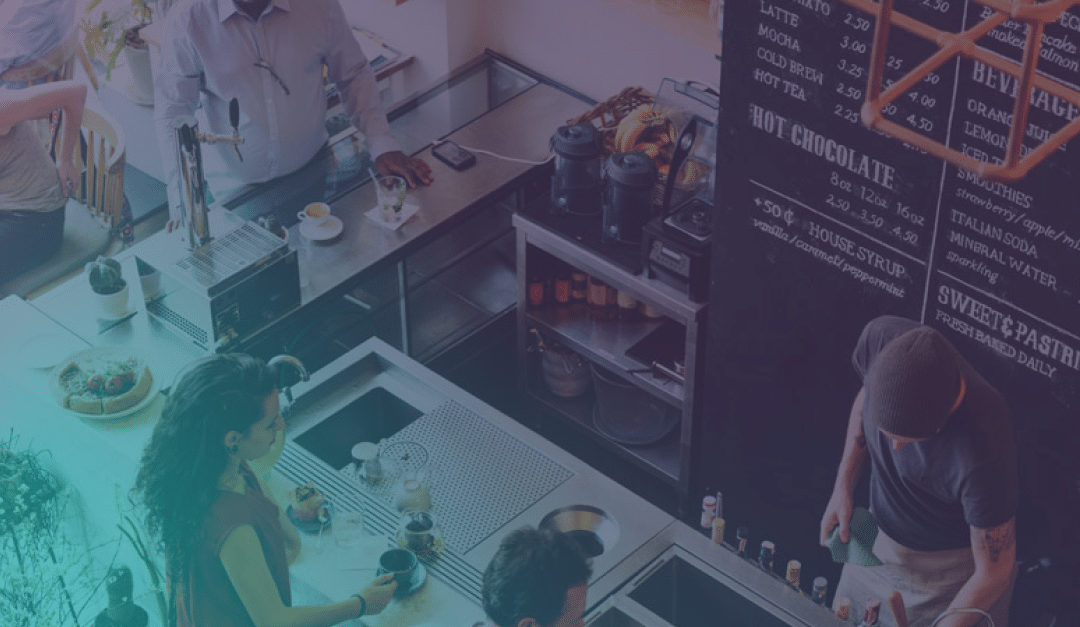 Vista aérea de la barra de una cafetería en la que se ve a gente pidiendo bebidas, a un barista preparando café y el menú escrito en una pizarra colgada en la pared.
