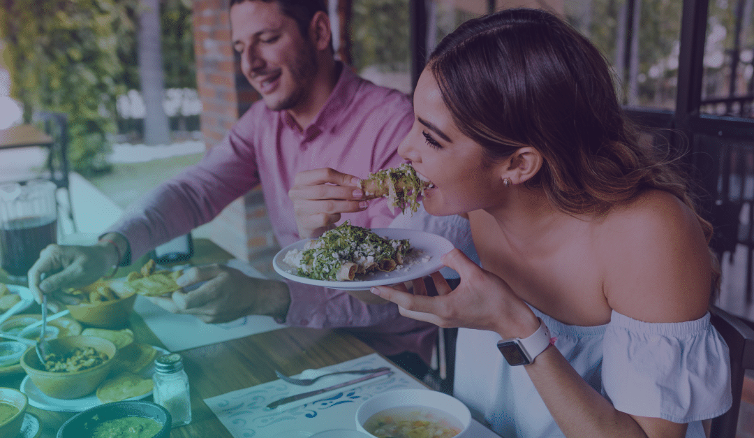 Una mujer y un hombre están sentados a una mesa, sonriendo y comiendo tacos, con varios platos y bebidas delante de ellos, en un restaurante luminoso e informal.