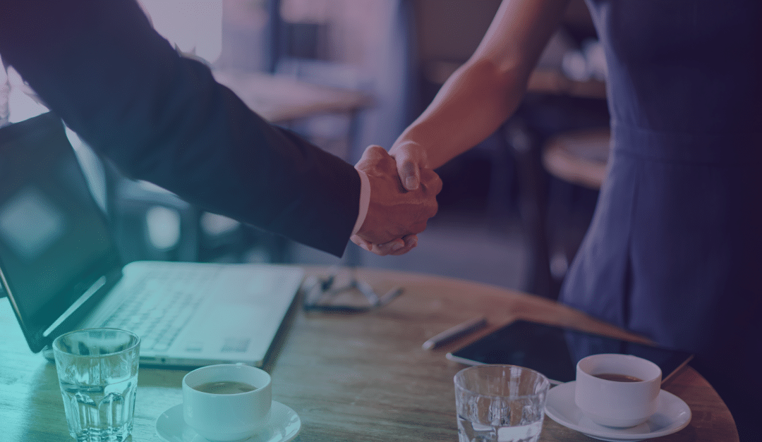 Dos personas vestidas con ropa de negocios dándose la mano al otro lado de una mesa en la que hay un ordenador portátil, una tableta y tazas de café, lo que sugiere una reunión profesional o un acuerdo.