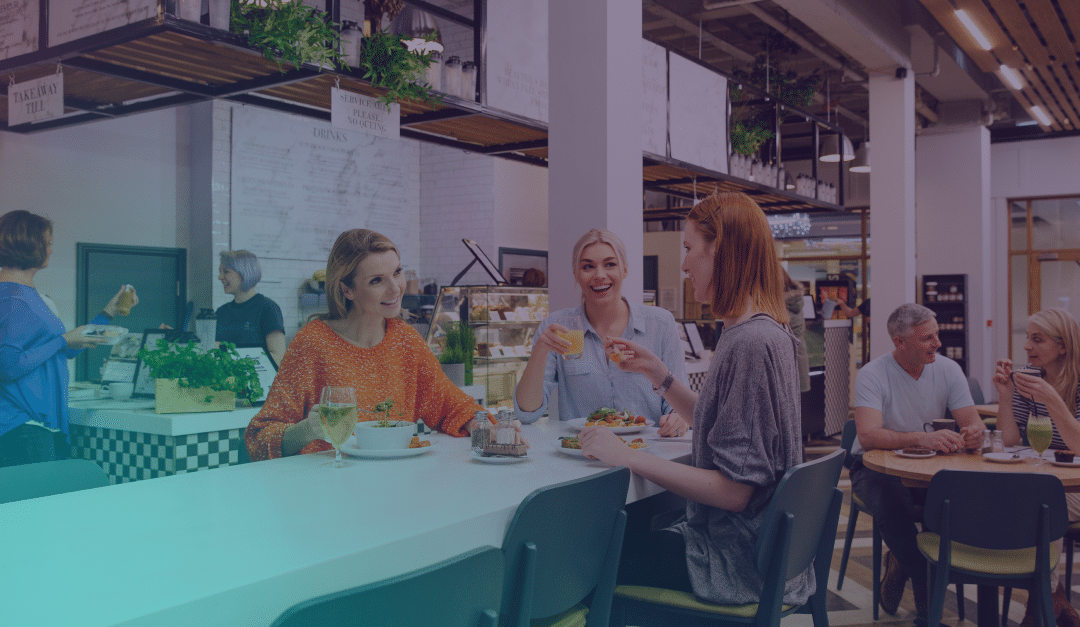Tres mujeres están sentadas en la barra de un restaurante moderno, sonriendo y comiendo juntas. Al fondo se ve a otros comensales y al personal, y el local cuenta con plantas y una iluminación brillante.