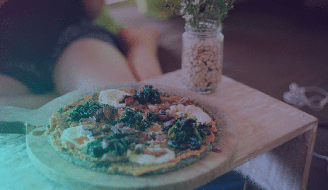 Una pizza con verduras, champiñones y queso reposa sobre una tabla de madera. Detrás de ella hay un tarro de alubias y una persona tumbada, difuminada, con algo de vegetación y unos auriculares al fondo.