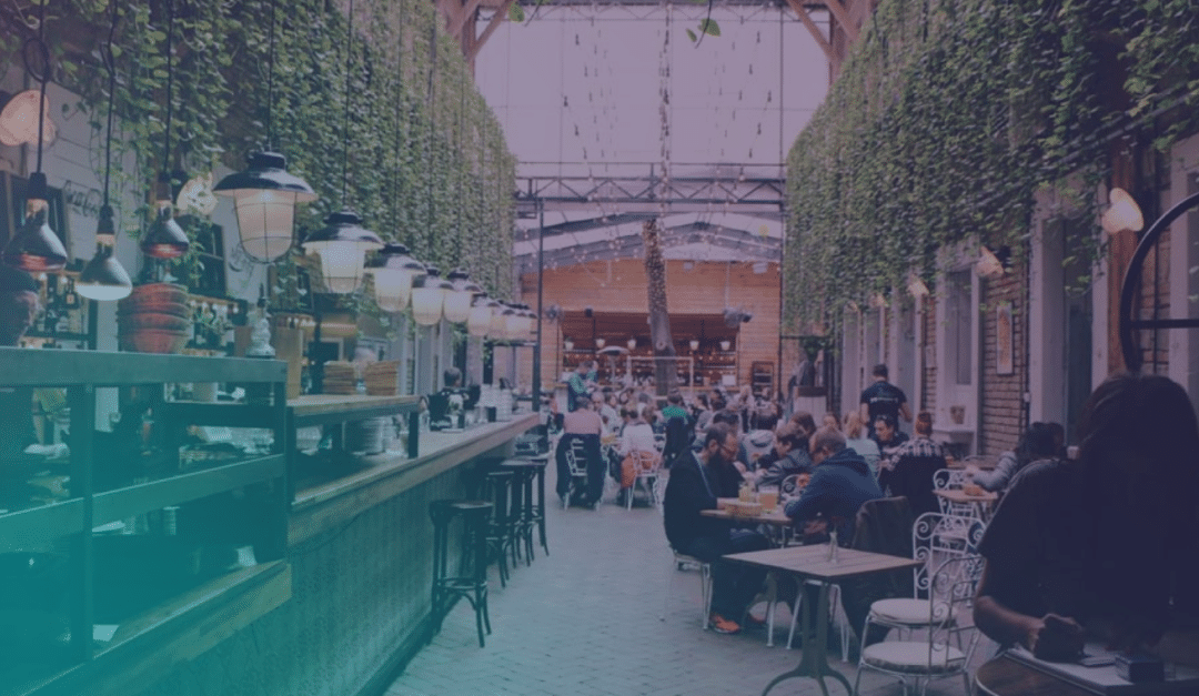 Gente cenando y charlando en un restaurante de estilo mercado cubierto, con plantas colgantes, guirnaldas de luces y un ambiente acogedor y animado.