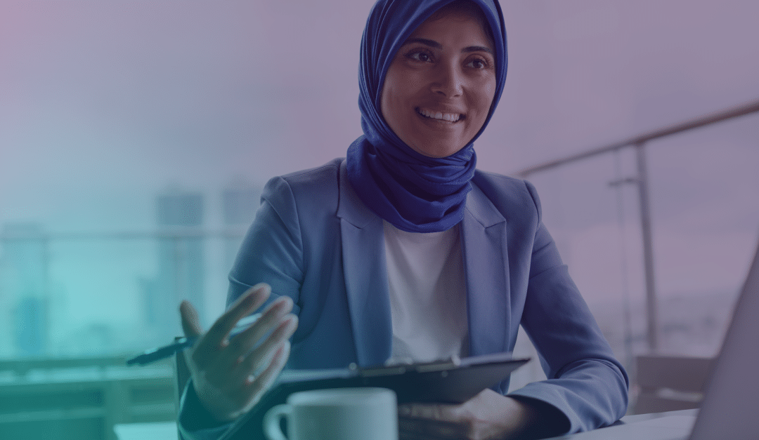 Una mujer con un hiyab azul y una chaqueta de traje está sentada ante un escritorio, sonriendo y sosteniendo una tableta con un lápiz. Sobre la mesa hay una taza de café y un ordenador portátil; al fondo, a través de unos grandes ventanales, se divisa un paisaje urbano difuminado.