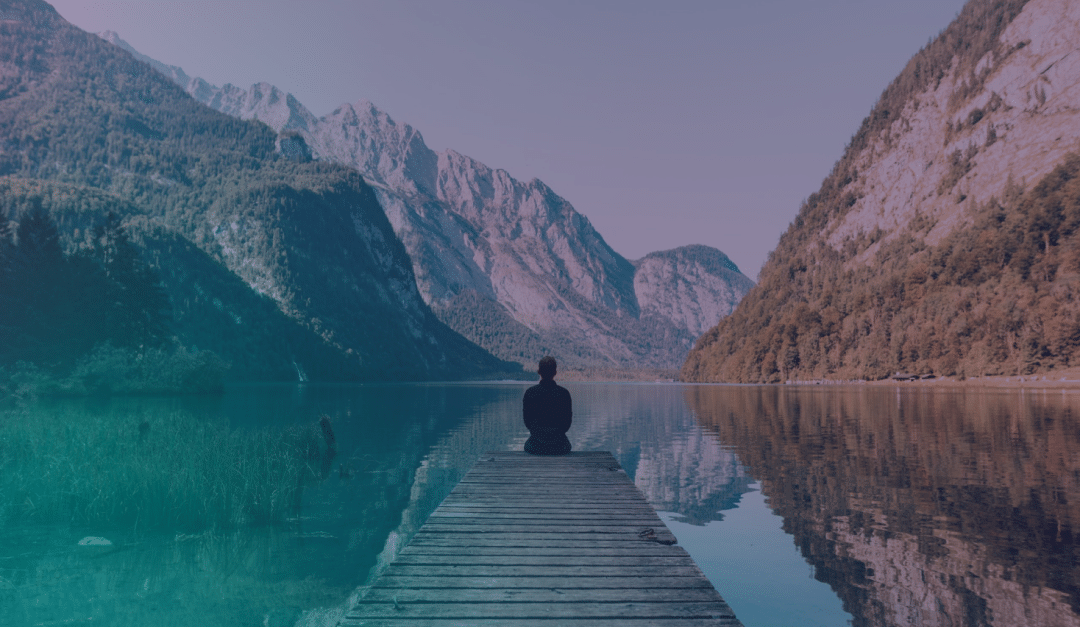 Una persona está sentada al final de un embarcadero de madera con vistas a un tranquilo lago de montaña, rodeada de picos altos y escarpados y laderas boscosas, bajo un cielo despejado.