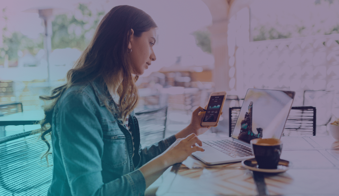 Una mujer está sentada en la mesa de una cafetería al aire libre, utilizando un ordenador portátil y con un smartphone en la mano. Junto a ella, sobre la mesa, hay una taza de café. El ambiente es luminoso y relajado.