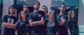 Un grupo de siete jóvenes vestidos con camisetas negras está de pie junto a un edificio, algunos con los brazos cruzados, mirando con seguridad a la cámara.