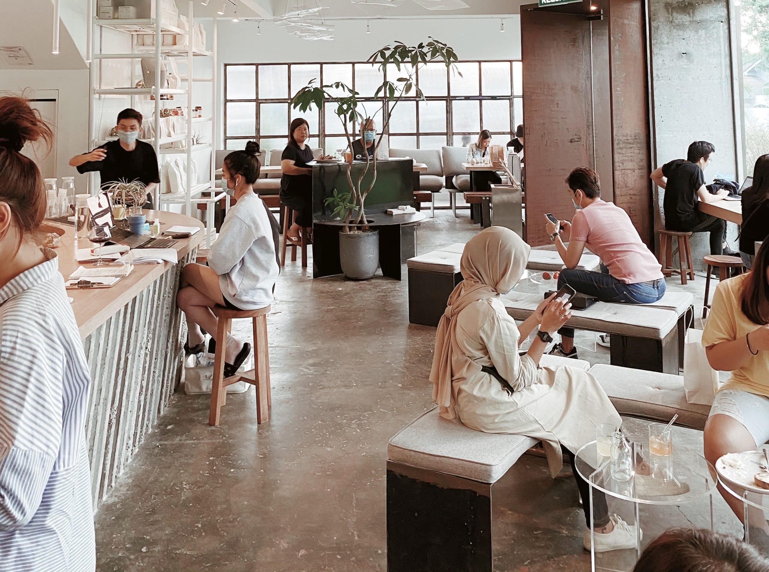 Una cafetería moderna en la que hay gente sentada en mesas y bancos; algunos charlan, otros usan el móvil. El personal sirve bebidas y los grandes ventanales dejan entrar la luz natural. Las plantas y la decoración minimalista crean un ambiente relajado.