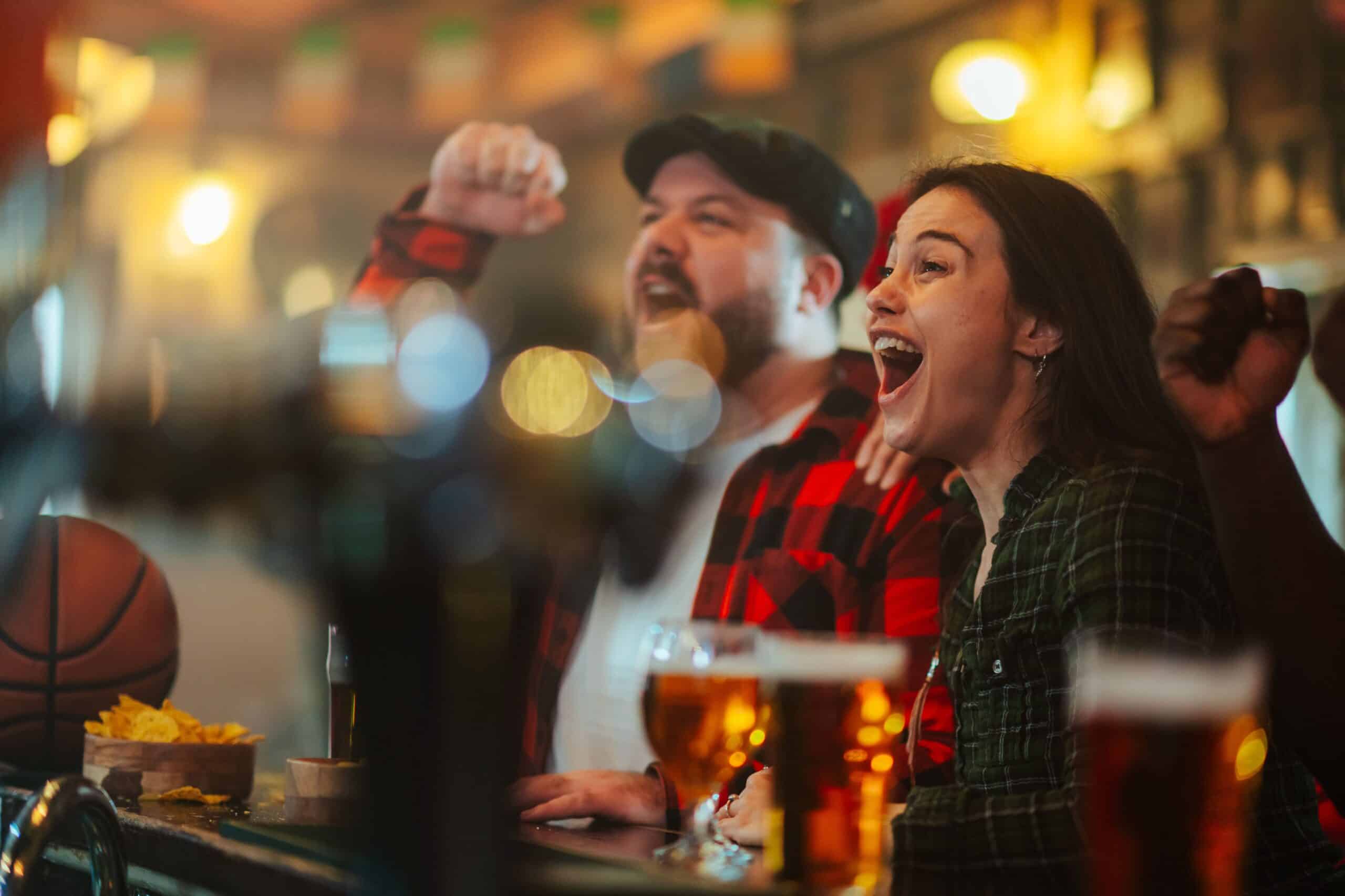 Dos personas en un bar vitorean con entusiasmo mientras observan algo que queda fuera de plano. Están rodeadas de bebidas y aperitivos, en un ambiente festivo y con luces difuminadas al fondo.