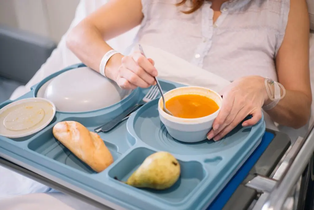 Una persona tumbada en una cama de hospital come sopa de un cuenco que hay sobre una bandeja azul, junto a la cual hay pan, una pera y otros platos tapados. La persona lleva una bata de hospital y una pulsera de identificación.
