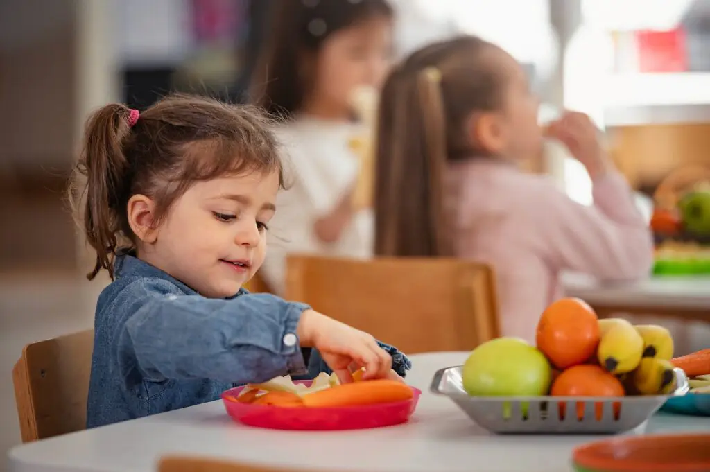 Una niña está sentada a una mesa cogiendo comida de un plato, con varias frutas en una bandeja cerca de ella. En el fondo, otros niños están sentados a la misma mesa, comiendo y charlando.