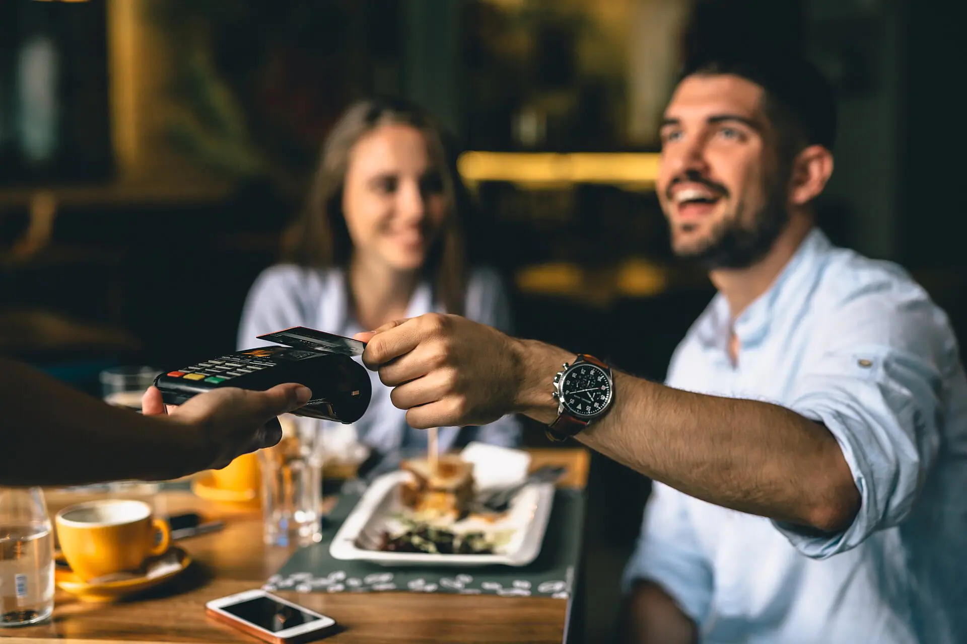 Un hombre sonriente le entrega una tarjeta de crédito a un camarero que sostiene un lector de tarjetas junto a una mesa de restaurante, con una mujer sentada a su lado y comida y bebidas sobre la mesa.