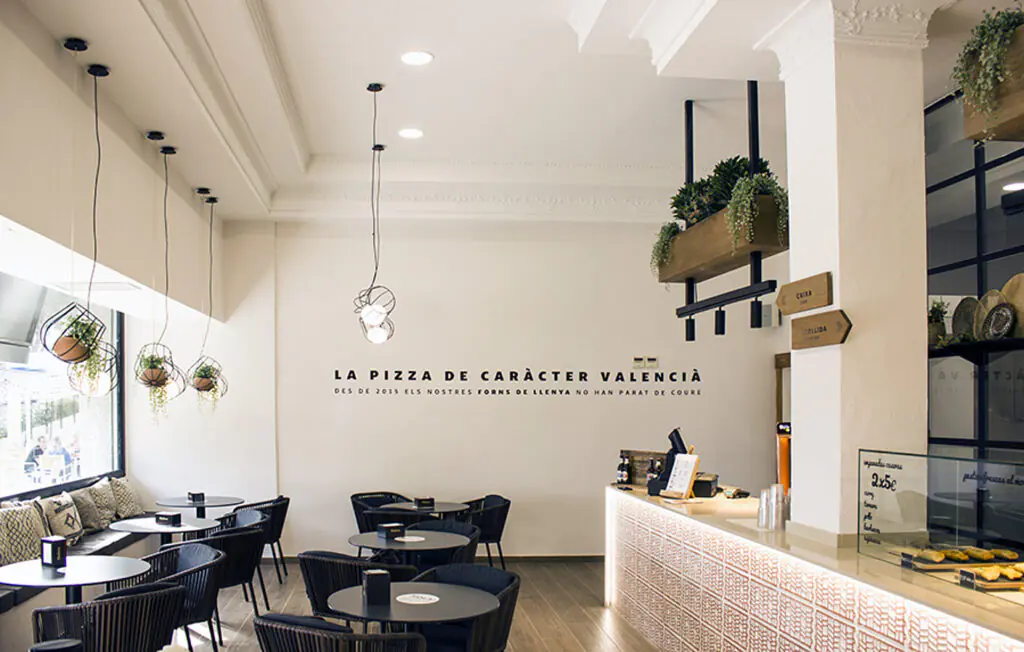 Una pizzería moderna y luminosa con sillas negras, mesas redondas, plantas colgantes y una barra en la que se exponen las pizzas. La pared blanca está decorada con texto en español.