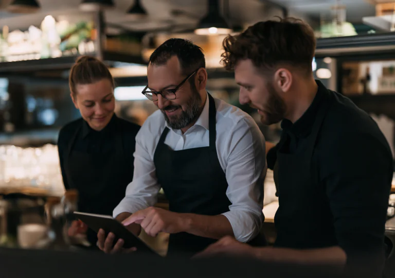 Tres personas con delantal están juntas en la cocina de un restaurante, sonriendo y mirando una tableta digital que una de ellas sostiene y utiliza, lo que sugiere un trabajo en equipo o una conversación sobre tareas laborales.