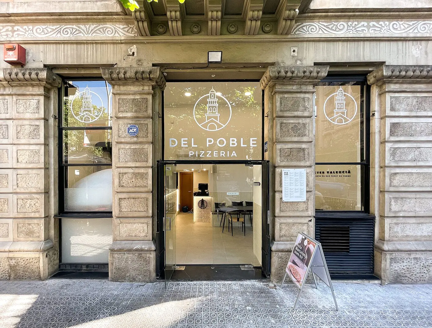 La entrada de la pizzería Del Poble, con grandes ventanales, una puerta de cristal y un expositor con la carta en el exterior. El logotipo y el nombre aparecen en los ventanales. El edificio tiene una fachada de piedra.