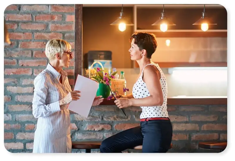 Dos mujeres mantienen una conversación seria en el interior, junto a una pared de ladrillo; una sostiene unos papeles y la otra, un ramo de flores, sentada en un taburete de bar. Al fondo se ven una iluminación cálida y unas estanterías.