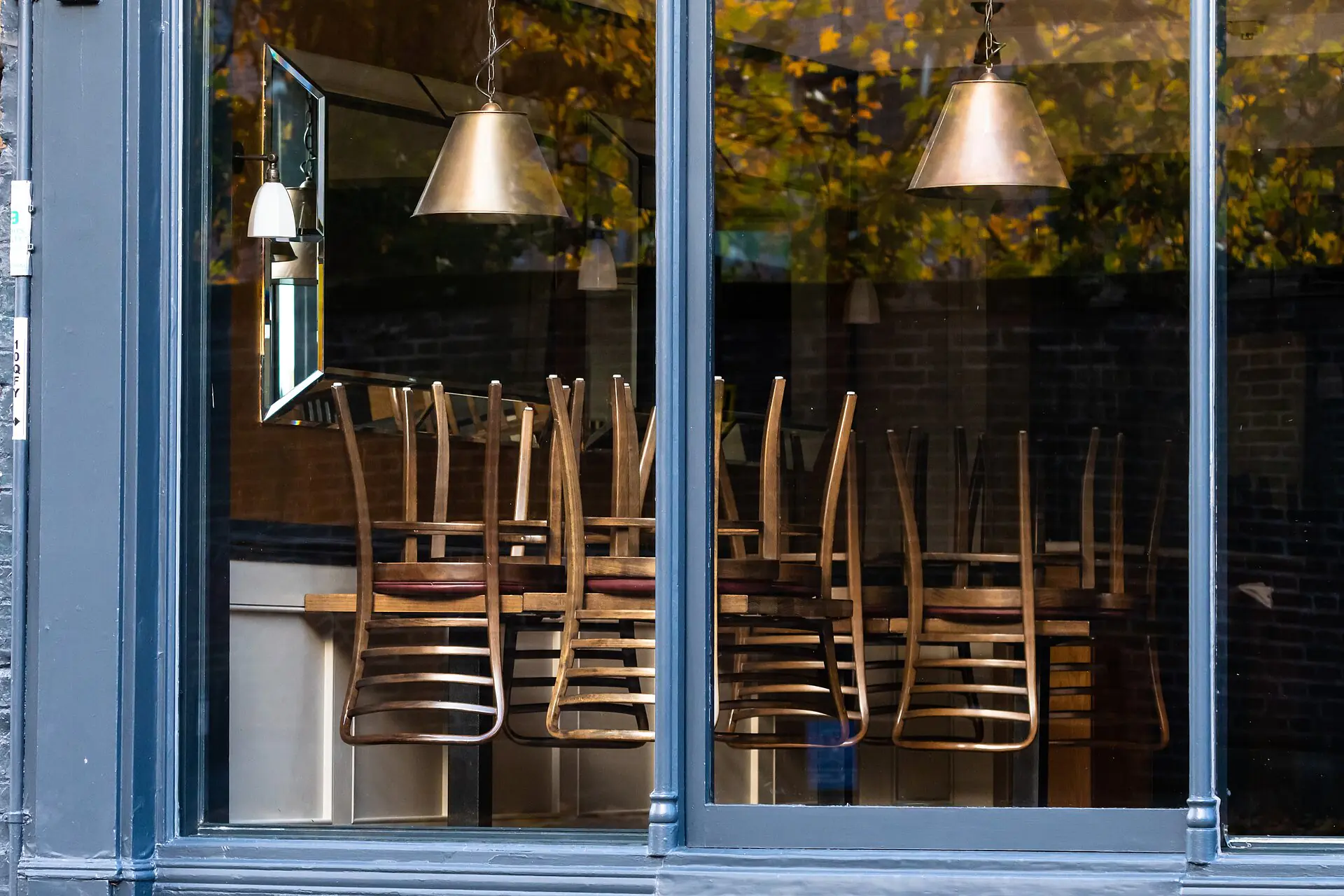 A través de un gran ventanal se ven sillas apiladas boca abajo sobre las mesas en el interior de un restaurante vacío y cerrado, con lámparas colgantes y los reflejos de los árboles del exterior.