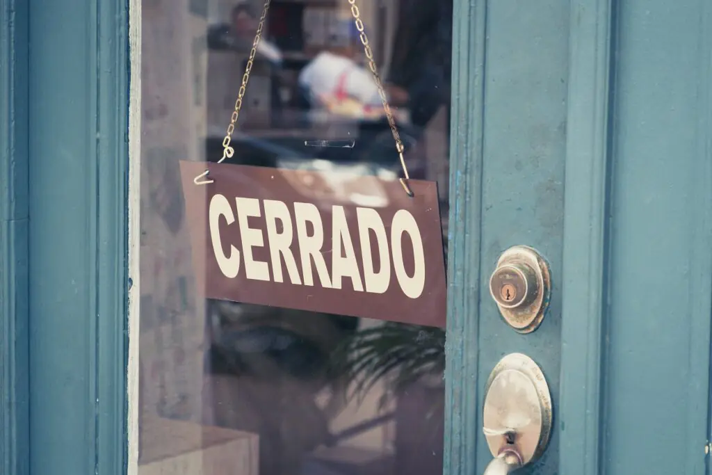 En una puerta de cristal cuelga un cartel marrón con la palabra «CERRADO», lo que indica que el local está cerrado. La puerta es de color verde azulado y se ve un cerrojo plateado.