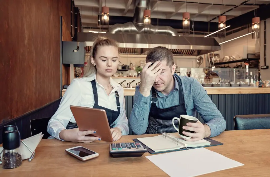 Dos empleados de una cafetería están sentados en una mesa con una tableta, una calculadora y unos papeles. La mujer mira la tableta, mientras que el hombre, que sostiene una taza de café, parece estresado y se lleva la mano a la frente.