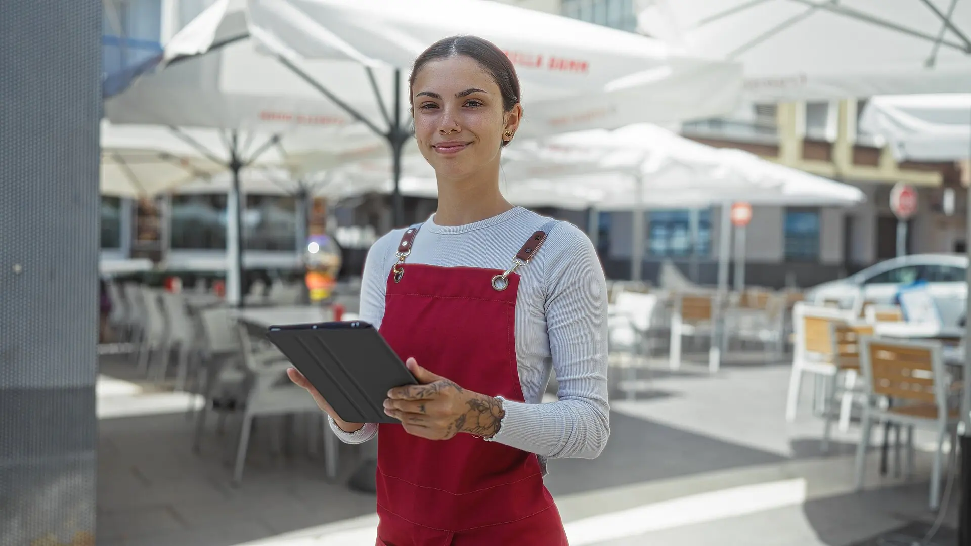 Una mujer con un mono rojo está de pie a la puerta de una cafetería, con una tableta en la mano y sonriendo. A su alrededor hay sombrillas blancas y mesas vacías en un día soleado.