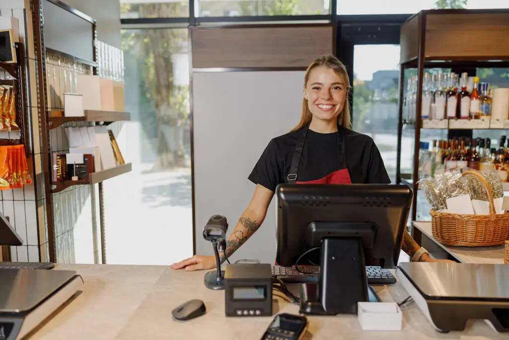 Una cajera sonriente se encuentra detrás del mostrador de una tienda bien iluminada, rodeada de estanterías con botellas, aperitivos y una cesta de pan, lista para atender a los clientes.