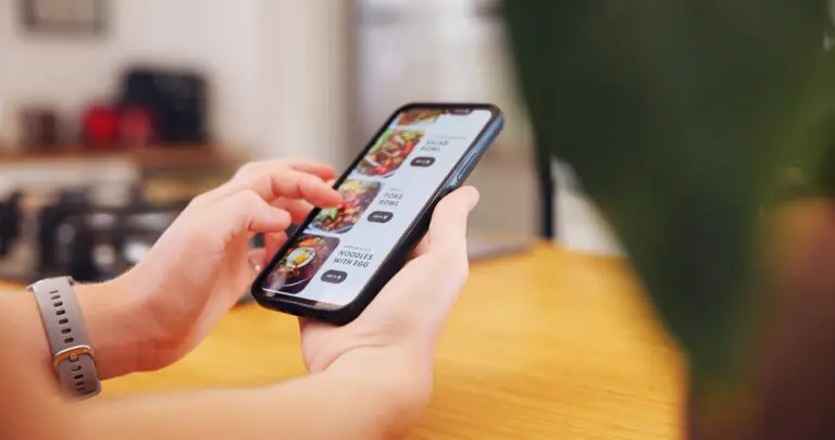 Una persona sostiene un smartphone y navega por una aplicación de reparto de comida a domicilio, seleccionando entre las opciones de menú. La escena se desarrolla en el interior y, en primer plano, se ve parte de una planta.