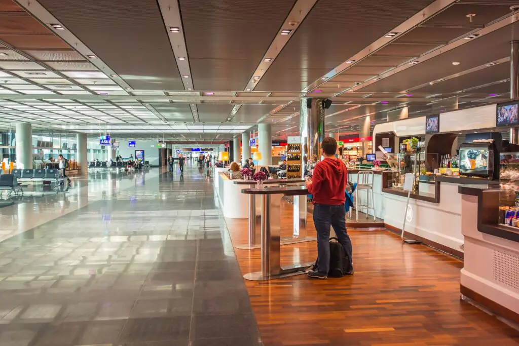 Una persona con una sudadera roja con capucha está de pie junto a la barra de una cafetería en la terminal de un aeropuerto; al fondo, bajo una luz intensa, se ven las zonas de descanso y las puertas de embarque.