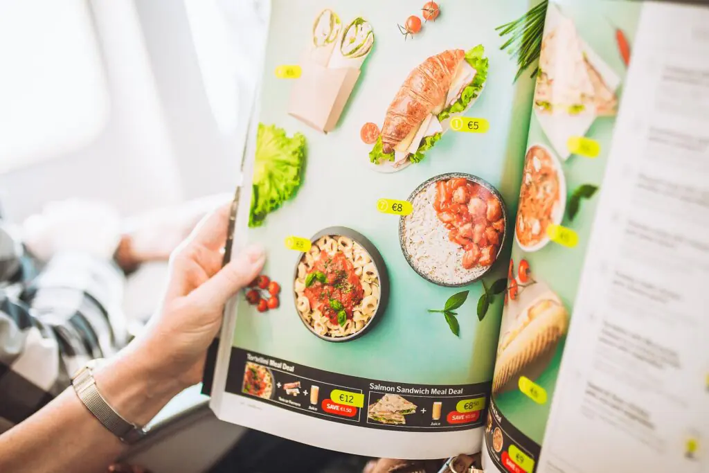 Una persona sostiene un menú de una compañía aérea en el que se muestran fotos a todo color de diversas opciones de comida con sus precios, entre las que se incluyen bocadillos, wraps, pasta, arroz y ensaladas, sobre un fondo verde claro.