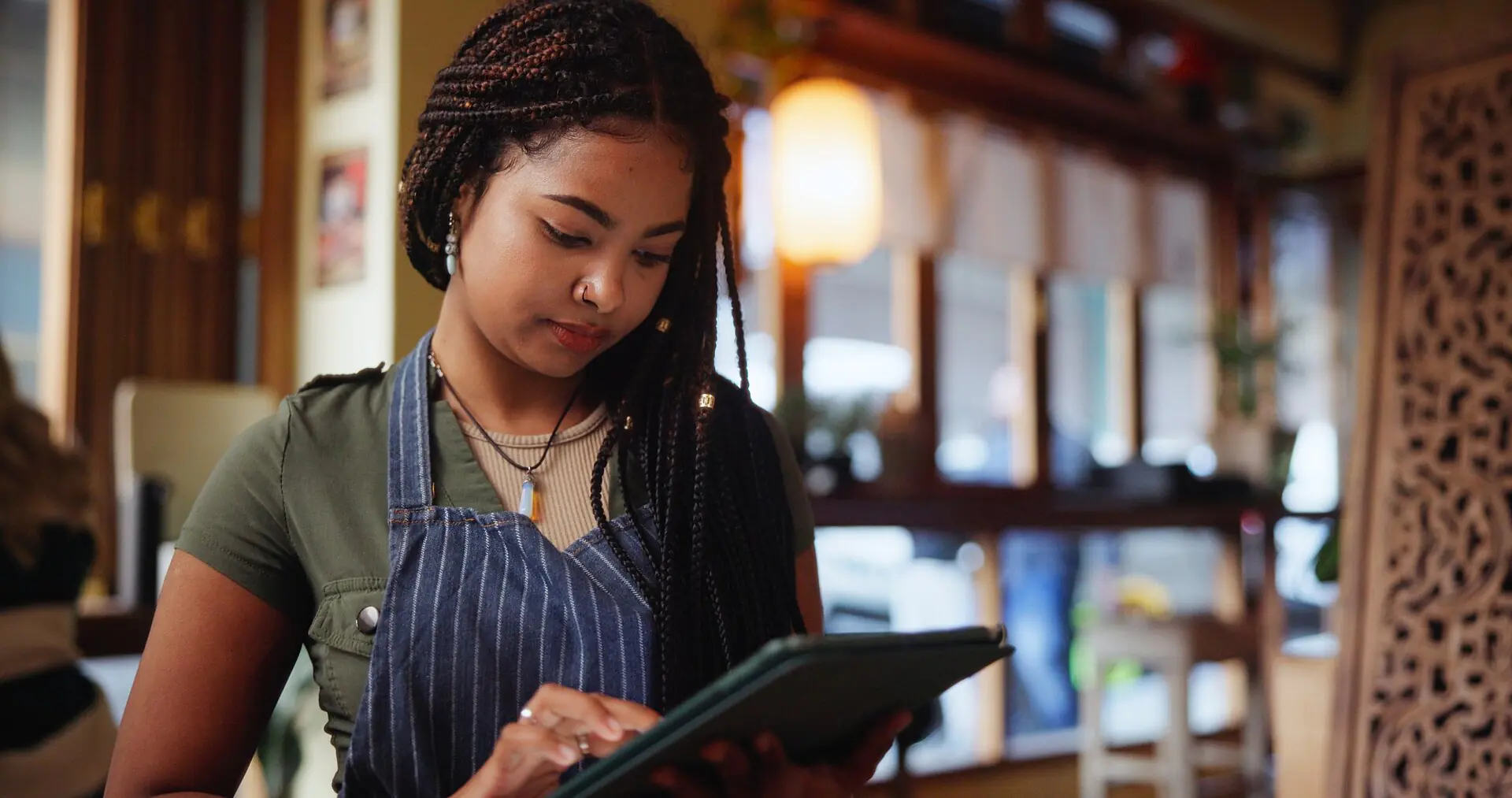 Una joven con un delantal a rayas utiliza una tableta en una acogedora cafetería con una iluminación cálida y una decoración de madera.
