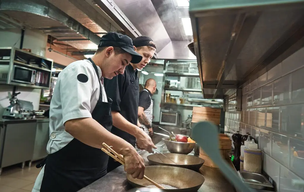 Dos cocineros, vestidos con uniforme, preparan comida en una cocina industrial, mezclando ingredientes en grandes cuencos metálicos sobre una encimera, con el equipamiento y los utensilios de cocina visibles al fondo.