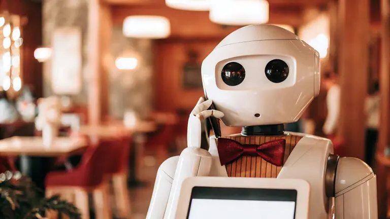 Un robot humanoide con ojos grandes, una pajarita roja y una pantalla táctil se lleva un teléfono a la oreja en un restaurante con una iluminación cálida, sillas rojas y decoración de madera.
