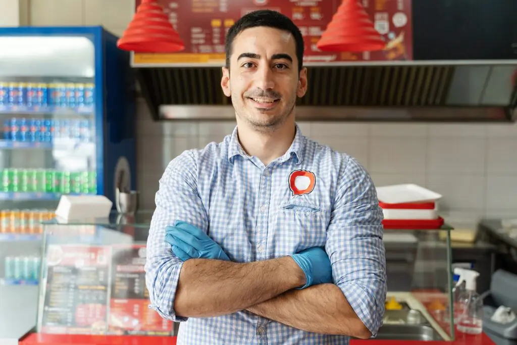 Un hombre que lleva una camisa a cuadros y guantes azules está de pie, con los brazos cruzados, en un restaurante de comida rápida, sonriendo con seguridad. Al fondo se ven las neveras de comida y bebida y un cartel con el menú.