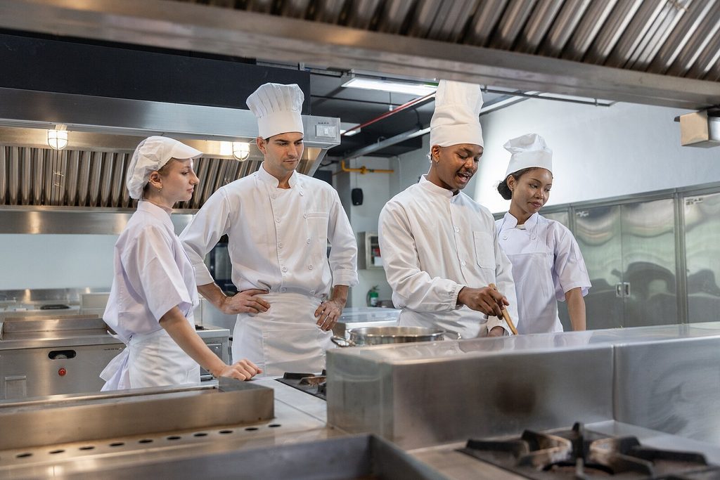 Cuatro cocineros, vestidos con uniformes blancos y gorros de chef, se encuentran en una cocina profesional; uno de ellos está explicando o haciendo una demostración a los demás junto a una cocina.